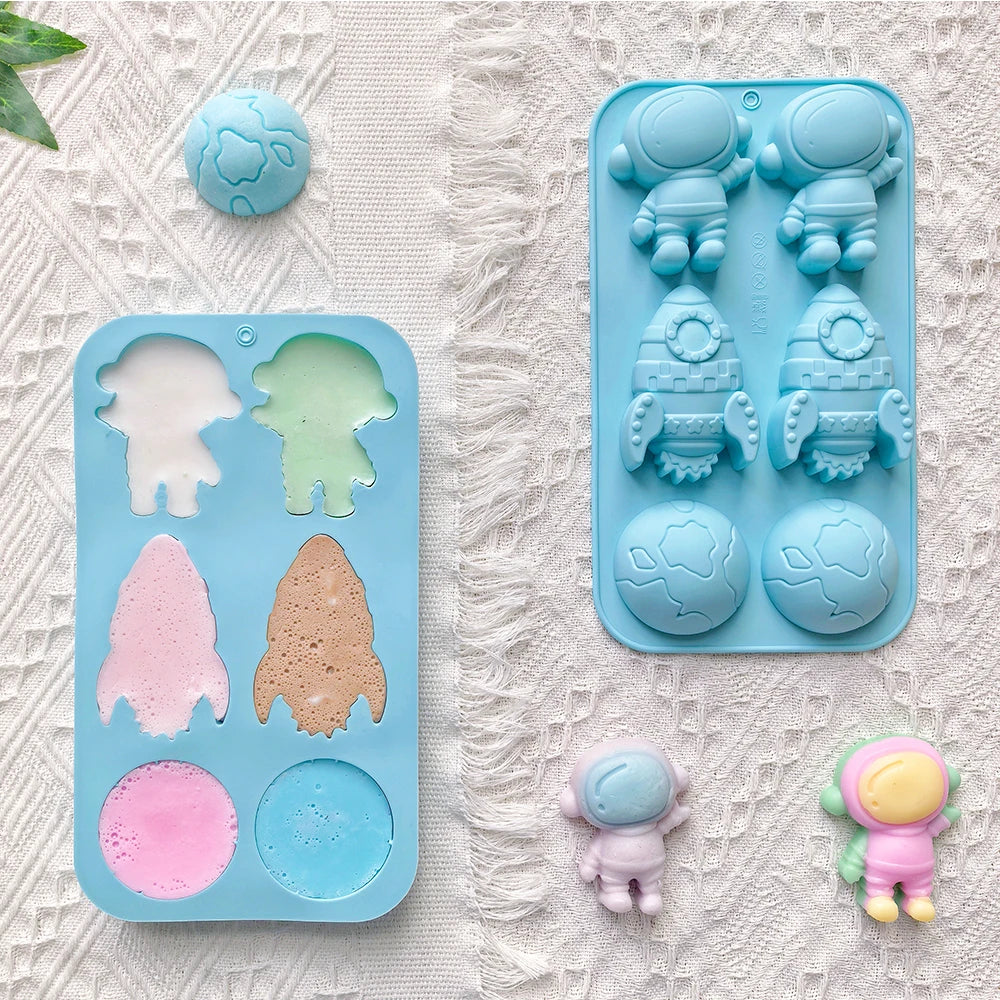 A silicone mold set for making astronaut-shaped ice pops or soaps, shown empty and filled with pastel colors on a textured white fabric background. The set includes a rectangular mold with astronaut and planet shapes, a round mold, and two small finished astronaut pieces.